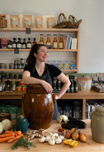 Load image into Gallery viewer, Expert fermenter Caitlin Walsh stands behind a wooden counter in a shop, smiling while leaning on a large brown ceramic jar. The counter is styled with fresh ingredients including carrots, garlic, ginger, onions, and oranges.
